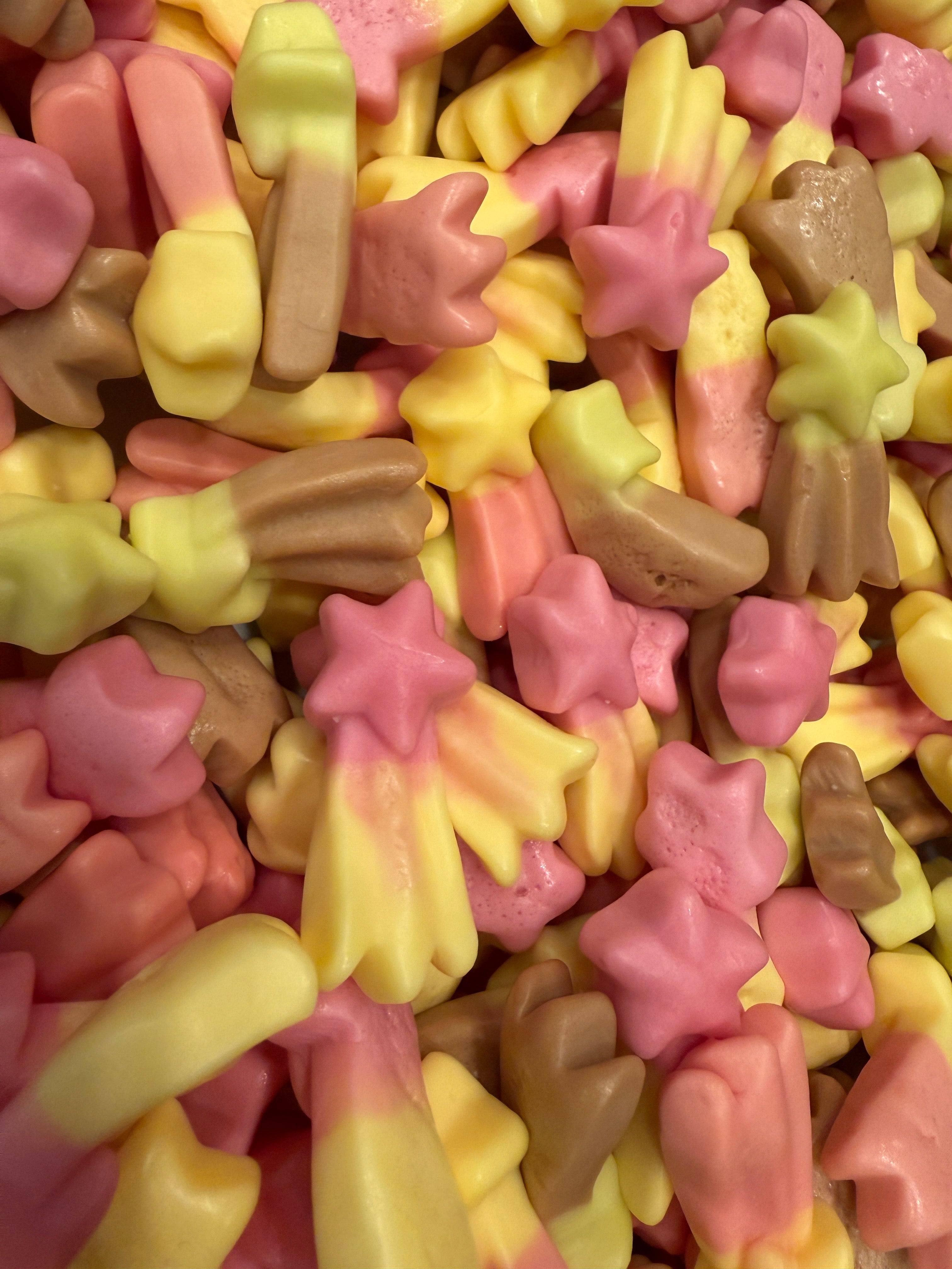 A close-up pile of small candy pieces shaped like stars, flowers, and abstract blobs in pastel pink, pale yellow, and light brown tones.