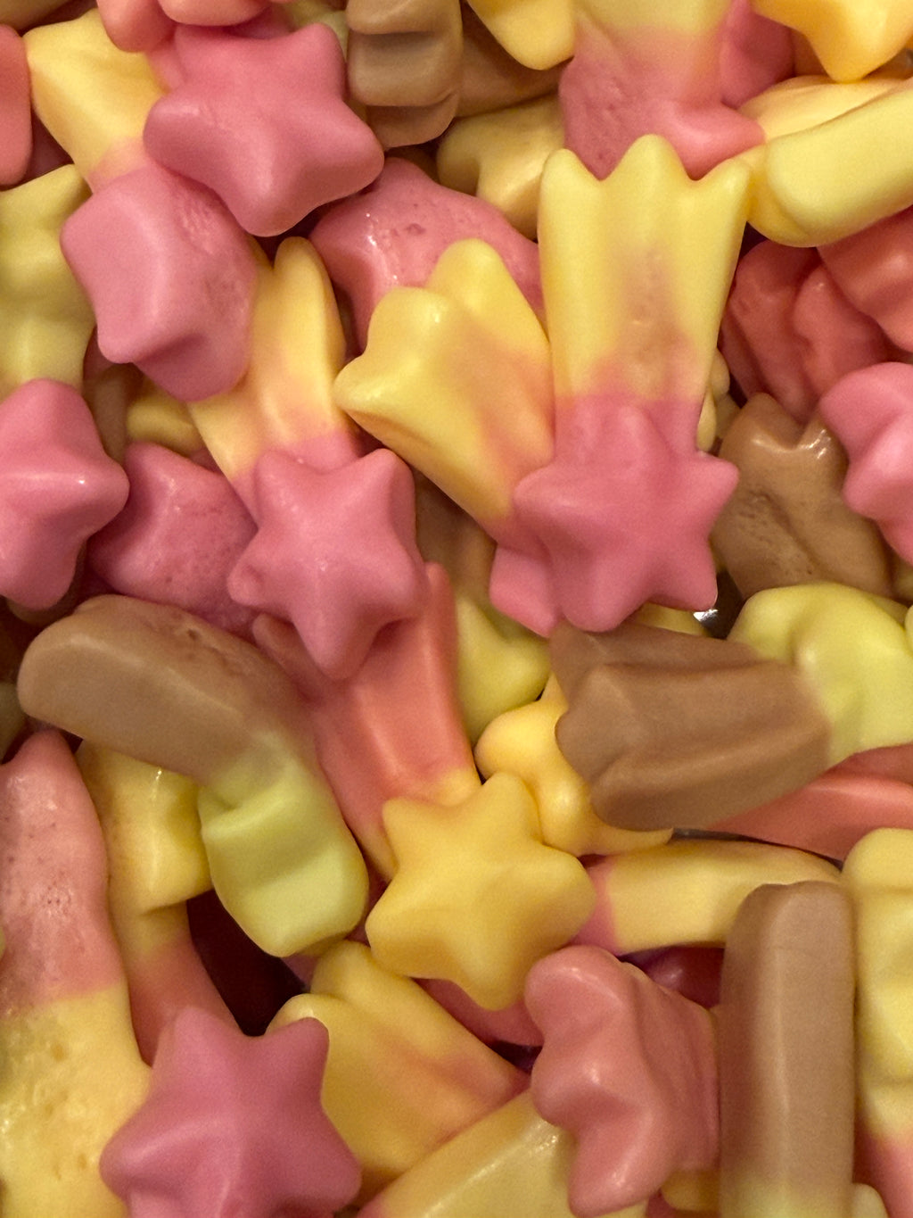 Extreme close-up of pastel star-shaped and elongated shooting-star gummies in pink, yellow, and light brown, with fewer, larger pieces clearly visible.