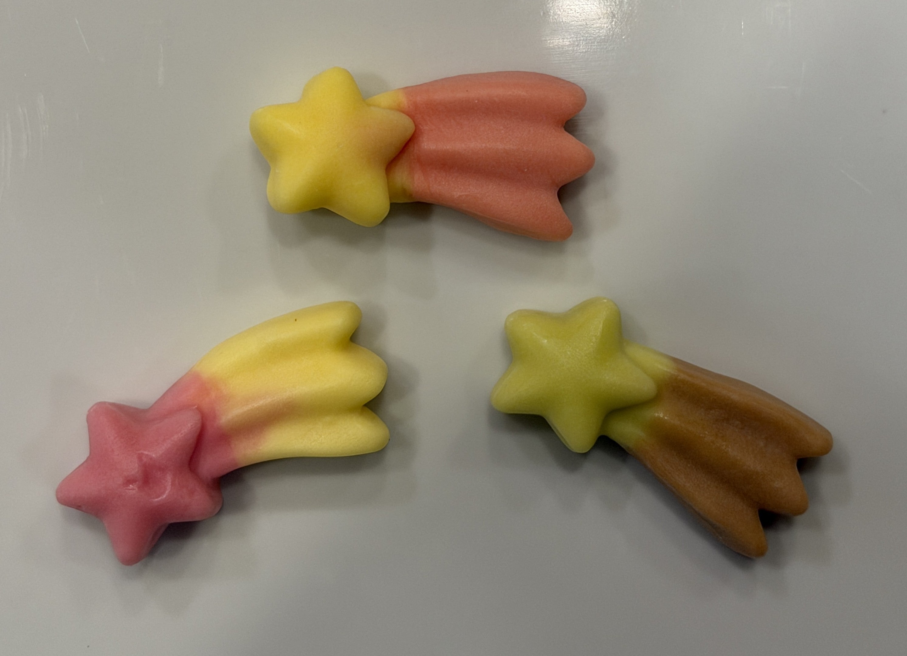 Three shooting star-shaped gummy candies in pink, yellow, peach, and brown, arranged on a plain light gray surface
