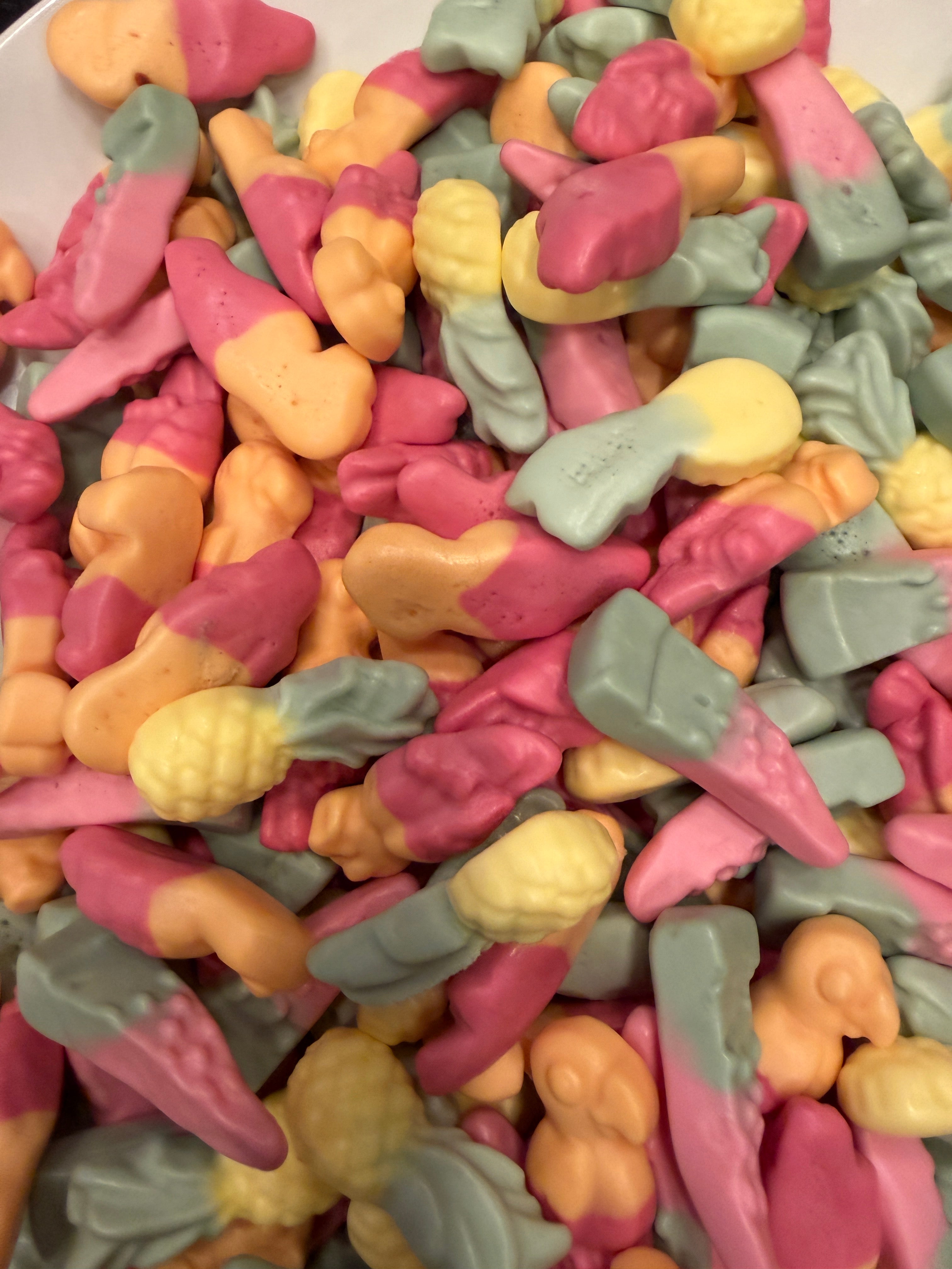 A close-up pile of colorful tropical fruit-shaped squishy candy pieces in pink, orange, pale yellow, and light gray-green, filling the entire frame.