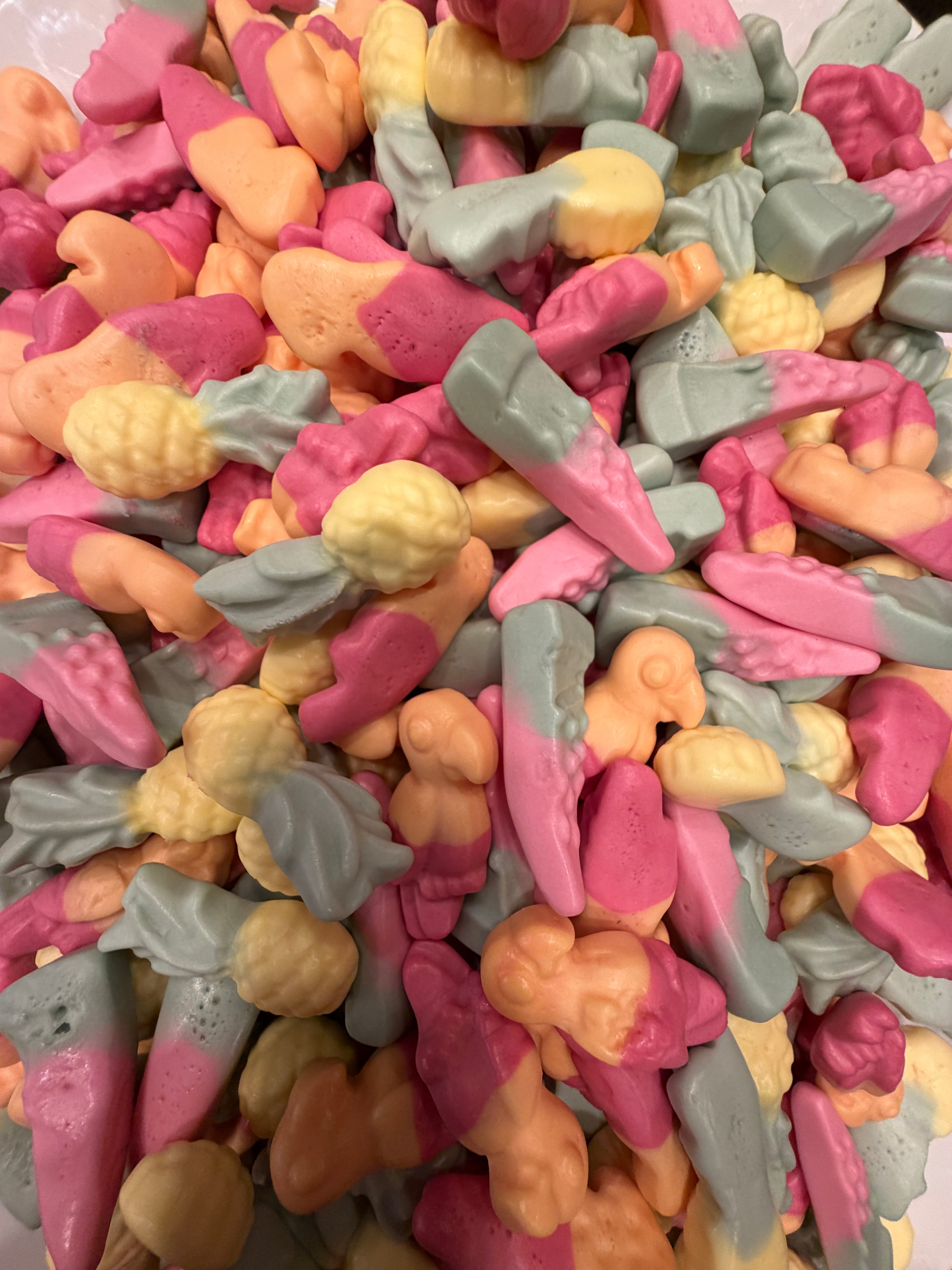 A close-up pile of small, colorful candy-like pieces shaped like vegetables and fruits, including carrots, pumpkins, and leaves, in pink, peach, yellow, and pale blue-green.