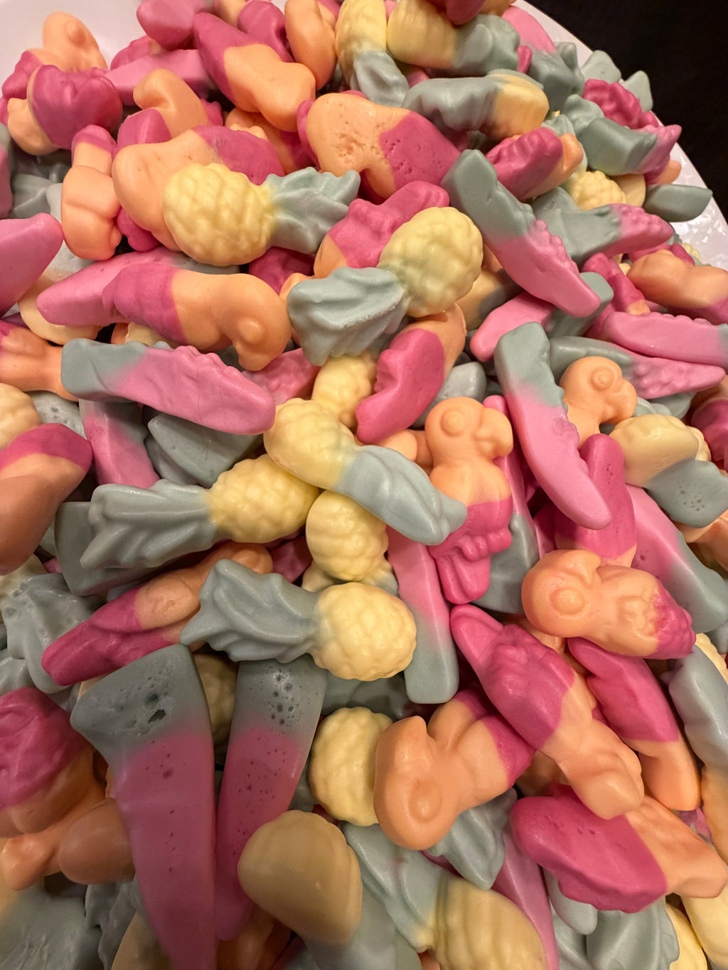 A close-up pile of colorful novelty gummy candies shaped like small figures and objects, in pink, peach, pale yellow, and light blue-gray tones.