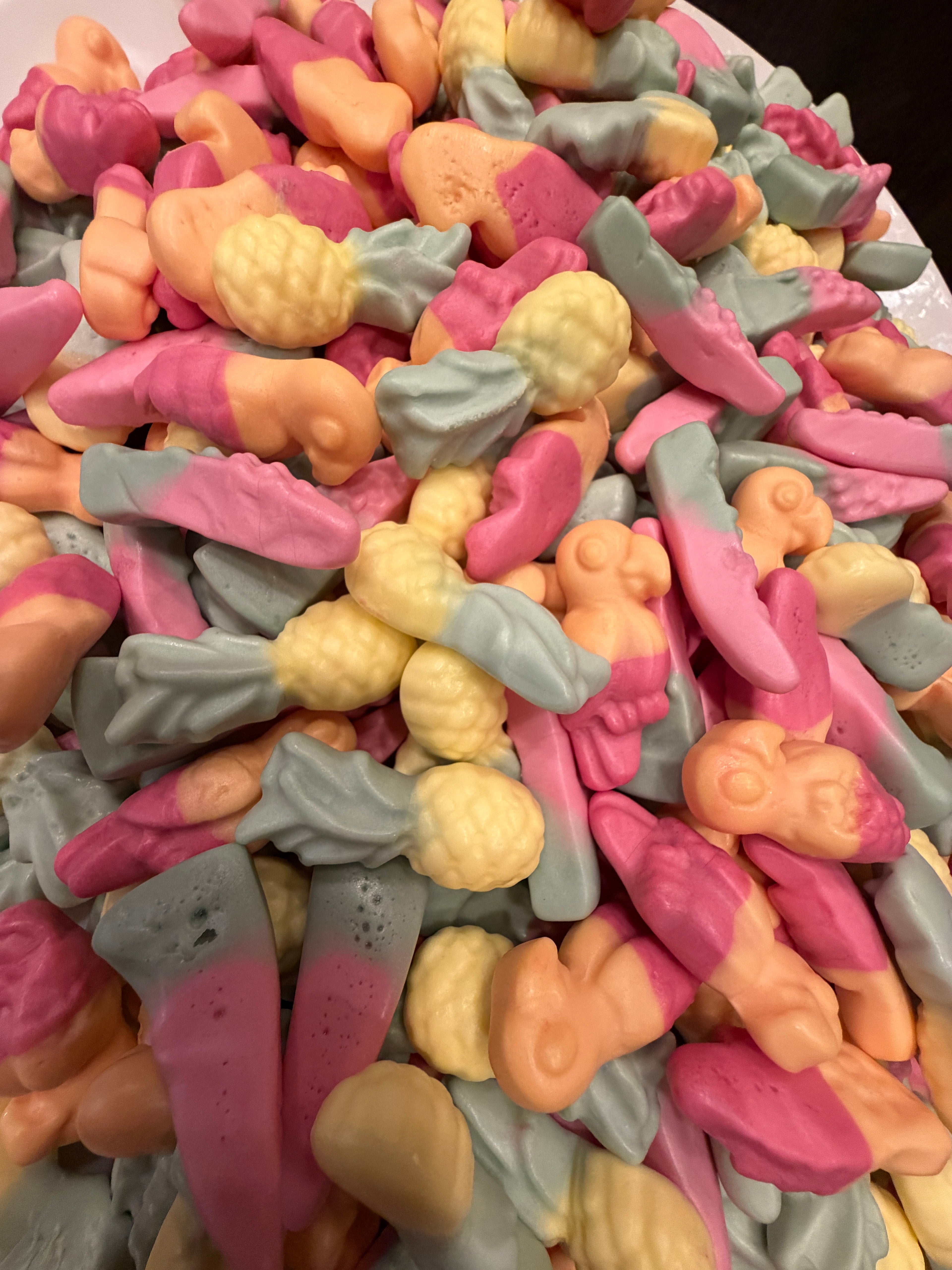 A close-up pile of colorful novelty gummy candies shaped like small figures and objects, in pink, peach, pale yellow, and light blue-gray tones.