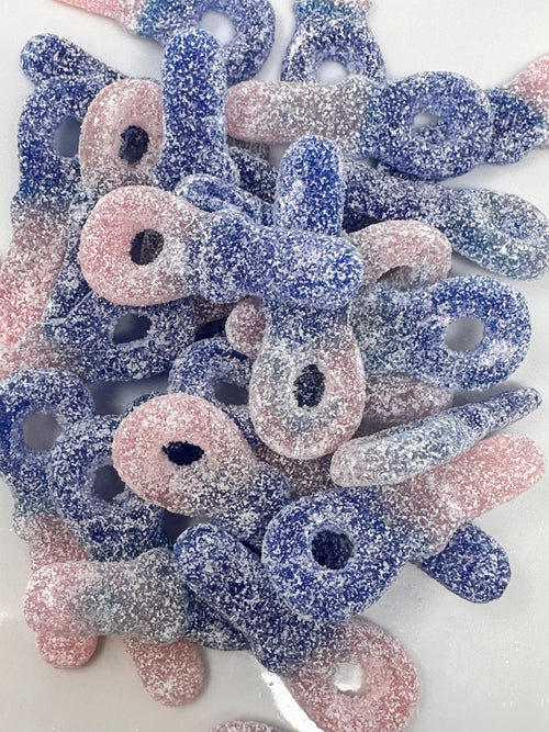 Overhead view of a pile of sugar-coated blue and pink pacifier-shaped gummy candies on a white surface, showing full candy pieces with open loop ends.